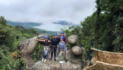Four people wearing helmets and harnesses stand on a rock formation surrounded by lush greenery, with a lake visible in the background.