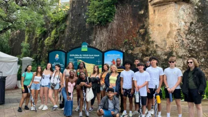 A group of young people stand in front of a sign and a historical wall, posing for a photo. Some hold water bottles and backpacks.