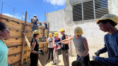 A group of people wearing construction helmets work together, passing buckets near a wooden structure under a sunny sky.