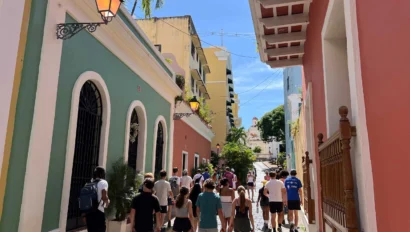 People walking down a narrow street lined with colorful buildings, under a clear blue sky.