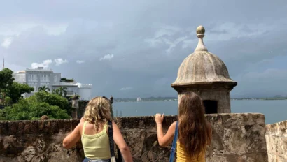 Two women with bags stand next to a historic stone building overlooking a body of water under a cloudy sky.