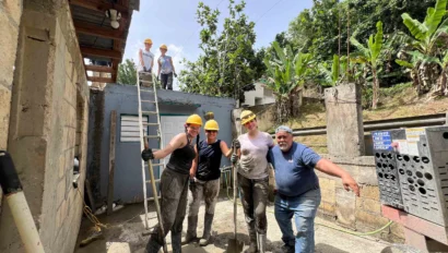 A group of people wearing hard hats and work clothes stand and pose while doing construction work in a partially built structure surrounded by trees.