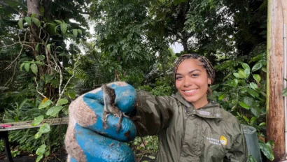 A participant on a teen service trip to Puerto Rico in a green jacket and blue gloves holds up a small lizard in a lush, tropical forest setting.