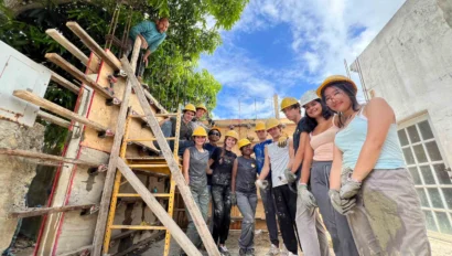 A group of teens yellow hard hats and work clothes pose at a construction site during a Puerto Rico hurricane relief service trip for teens, standing near a ladder and a partially built wall under a tree and blue sky.
