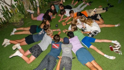 A group of people lying on the grass in a circle, focused on an object in the center.