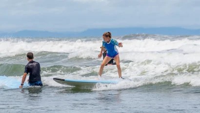 A child in a blue shirt practices surfing on a small wave, while an adult in the water offers assistance.