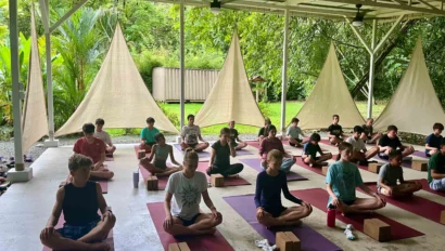 A group of people practice seated yoga indoors on mats, surrounded by green foliage and large fabric shades.