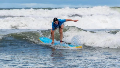 A person surfing on small waves with a blue surfboard, wearing a blue rash guard, and bending forward with an arm extended for balance.