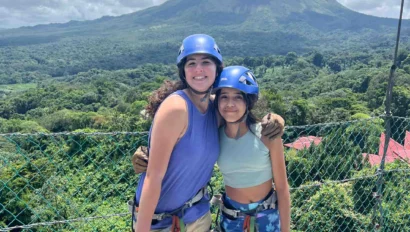 Two people wearing helmets and harnesses stand on a platform, ready for adventure as part of a Costa Rica Travel Program, with a lush green landscape and a large volcano in the background.