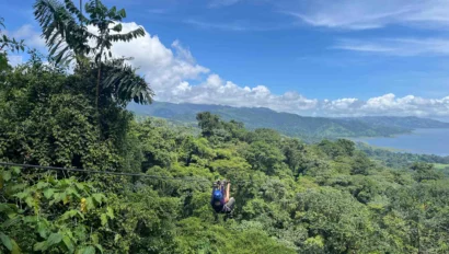 Person ziplining over a lush green rainforest with a view of distant mountains and a lake under a clear blue sky.