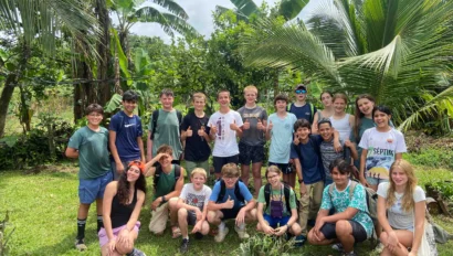 A group of teens poses outdoors in front of lush greenery with palm trees in the background. They are standing and crouching on a grass lawn, some smiling and giving thumbs up.