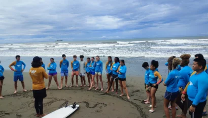 A group of people in blue shirts stand on a beach, forming a semicircle around an instructor. A surfboard lies in the sand. The ocean is in the background under a cloudy sky.