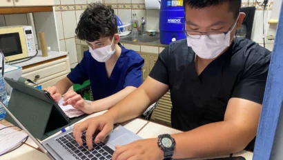 Two people wearing face masks work on a laptop and papers at a kitchen table, with a microwave and water cooler in the background.