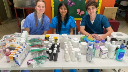 Three young people in scrubs sit behind a table filled with medical supplies and medications, in a brightly colored room with butterfly-themed wall decor, ready to share their expertise during a Dominican Republic school trip.