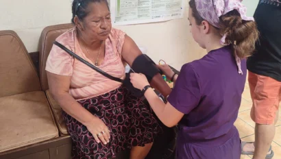 A healthcare worker in purple scrubs checks the blood pressure of a seated woman in a waiting area.
