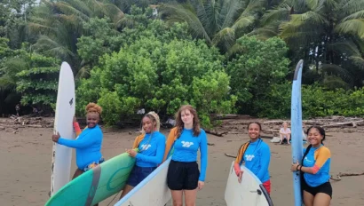 Five people in blue rash guards stand on a sandy beach holding surfboards, with palm trees and greenery in the background—capturing a moment from a Costa Rica student travel program under a cloudy sky.