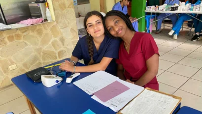Two women in scrubs sit at a blue table with medical forms, a blood pressure monitor, and clipboards, smiling at the camera in a clinic during a medical study abroad experience in Costa Rica.