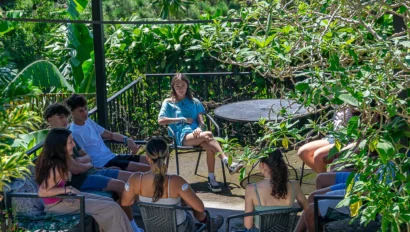 A group of people sits together outdoors on a patio surrounded by lush greenery, with one person sitting apart at a black table.