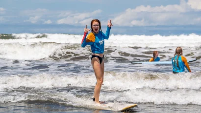 Person surfing on a wave, wearing a blue and orange wetsuit, making a peace sign with both hands.