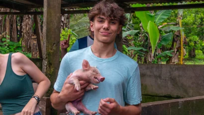 A young person in a blue shirt holds a piglet in an outdoor setting with greenery and a wooden structure in the background.
