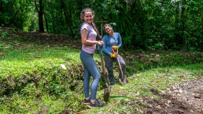 Two people in casual clothing are standing on a grassy path in a forested area, holding shovels and smiling at the camera.