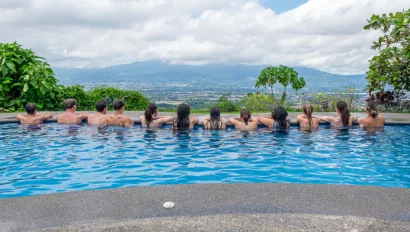 Eleven people sitting at the edge of a pool, overlooking a scenic landscape with mountains and cloudy sky in the background.