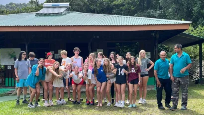 A group of people poses in front of a wooden building with a green roof surrounded by trees.
