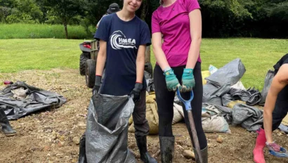 Two people in outdoor gear smile while holding a shovel and a sack, surrounded by bags and greenery.