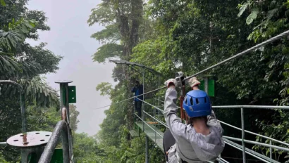 Person ziplining through a tropical forest canopy, wearing a blue helmet and gray jacket. Lush green trees surround the metal platform and zipline.