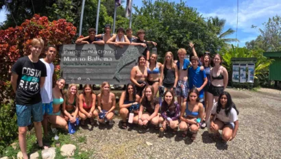 A group of people pose in front of the Marino Ballena National Park sign on a sunny day, surrounded by lush greenery and flags.