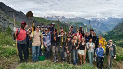 A group of people holding tools poses on a grassy hillside with mountains in the background under a cloudy sky.