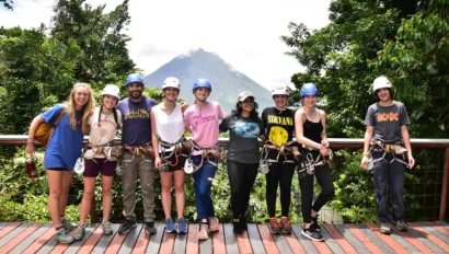 A group of eight people wearing helmets and harnesses stand on a wooden deck with a lush green forest and a mountain in the background.
