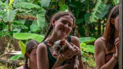 A young woman on a student travel to Costa Rica stands outdoors, holding a small piglet and smiling at the camera, with two other people and large green leaves in the background.