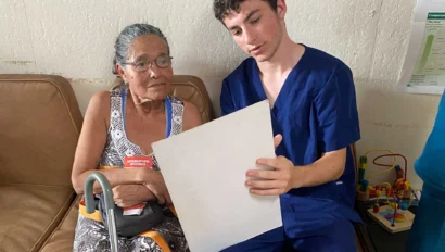A Global Works student in blue scrubs sits beside an older woman with a cane, showing her a document while they sit on a bench indoors—part of a Costa Rica student travel program focused on compassionate senior care.