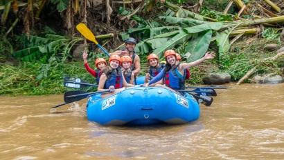 Six people wearing helmets and life jackets paddle a blue inflatable raft on a muddy river, smiling and waving at the camera with green foliage in the background—a thrilling moment from their Costa Rica student travel adventure.