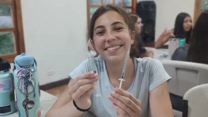 A young woman sits at a table holding two syringes, smiling at the camera during a school group travel event. An orange and several syringes are also on the table in front of her, with other people in the background.