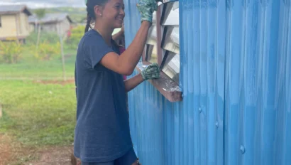 Person painting the exterior wall of a building blue with a brush, standing on grass.