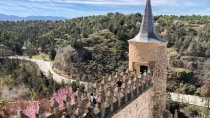 A medieval stone tower with a conical roof overlooks a lush, hilly landscape. People are visible on a walkway.