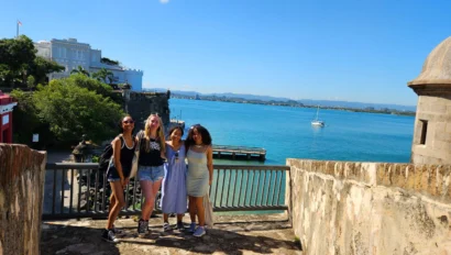 Four people stand smiling on a sunny day by a stone wall overlooking the ocean, with a sailboat in the water and buildings visible in the background.