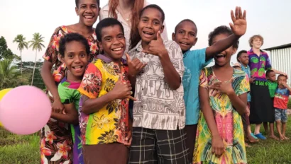 A group of smiling children and an adult stand together on grass, some giving thumbs up, with a pink balloon and trees in the background—capturing joy during a student travel program by Global Works.