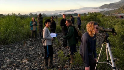A group of people is engaged in outdoor activities on a rocky terrain with binoculars and notes. Lush vegetation and mountains are visible in the background.