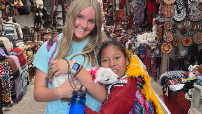Two people stand in a busy market filled with colorful textiles and crafts. One holds a white animal, and the other is smiling. Bright decorations hang from the ceiling.