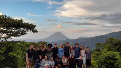 A group of people poses on grass with a volcano and lush greenery in the background under a cloudy sky.