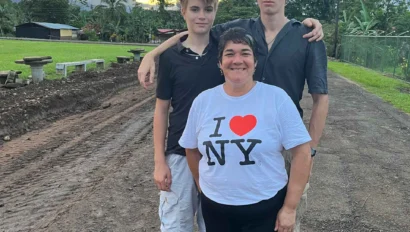 Three people stand on a dirt path with a sunset in the background. The person in the center wears an "I ♥︎ NY" shirt.