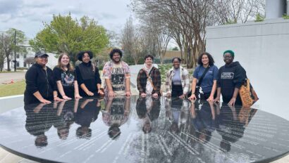 A group of nine people stands around a large, reflective, circular black table outside. Trees and an overcast sky are in the background.