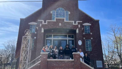 A group of people stands on the steps of a brick building under a clear blue sky.