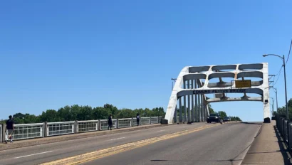 Edmund Pettus Bridge spans a clear blue sky with pedestrians on the walkway and a car driving across.