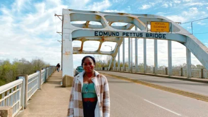 A woman stands on the sidewalk of the Edmund Pettus Bridge under a partly cloudy sky, reflecting on its legacy during a civil rights school trip.