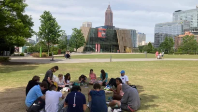 A group of people sitting in a circle on a lawn in an urban park, with modern buildings and trees in the background.