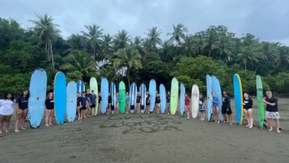 A group of people stands in a line on a beach, each holding a surfboard. "Woosh 2023" is written in the sand. Palm trees and cloudy sky are in the background.